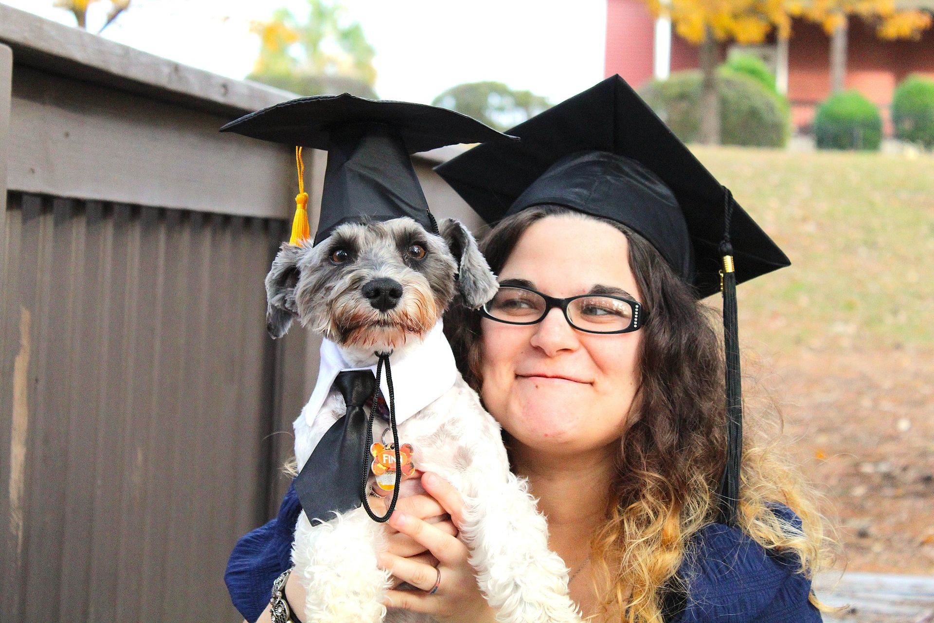 Woman and dog wearing graduation caps smiling outdoors.